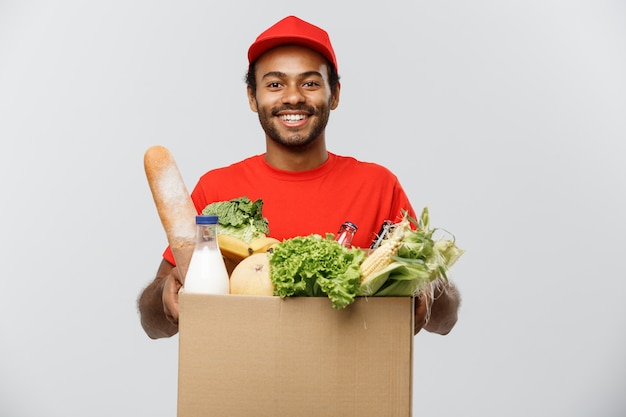 Concetto di consegna - bello uomo di consegna africano americano che trasporta scatola di pacchetti di alimentari e bevande al dettaglio da negozio. Isolato su sfondo grigio dello studio. Copia spazio.