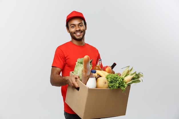 Concetto di consegna - bello uomo di consegna africano americano che trasporta scatola di pacchetti di alimentari e bevande al dettaglio da negozio. Isolato su sfondo grigio dello studio. Copia spazio.