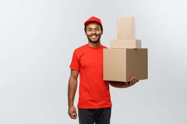 Concetto di consegna - Ritratto di felice uomo di consegna africano americano in panno rosso in possesso di un pacchetto di scatola. Isolato su sfondo grigio dello studio. Copia spazio.