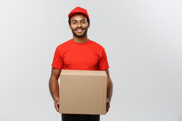 Concetto di consegna - Ritratto di felice uomo di consegna africano americano in panno rosso in possesso di un pacchetto di scatola. Isolato su sfondo grigio dello studio. Copia spazio.