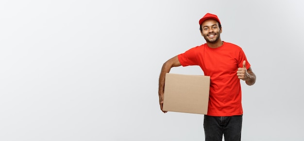 Concetto di consegna Ritratto di un uomo di consegna afroamericano felice che tiene un pacchetto di scatole e mostra i colpi isolati su uno spazio di copia di sfondo grigio dello studio