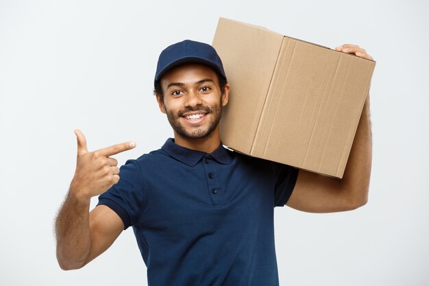 Concetto di consegna - Ritratto di uomo felice di consegna dell&#39;afroamericano che punta a presentare un pacchetto di scatola. Isolato su sfondo grigio dello studio. Copia spazio.