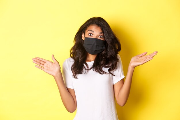 Concetto di coronavirus, pandemia e stile di vita. Immagine di una ragazza afroamericana carina in maschera facciale, che alza le spalle e sembra all'oscuro, non sa nulla, in piedi su sfondo giallo.