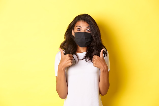 Concetto di coronavirus, pandemia e stile di vita. Ritratto di bella ragazza afro-americana in maschera nera, mostrando il pollice in su e guardando stupita, consigliando qualcosa, sfondo giallo.