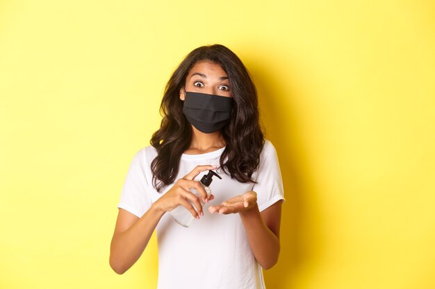 Concetto di covid-19, distanziamento sociale e stile di vita. Immagine della ragazza afro-americana in maschera facciale con disinfettante per le mani, in piedi su sfondo giallo.