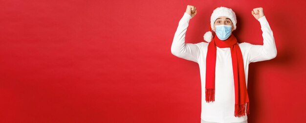 Concetto di covid-19, natale e vacanze durante la pandemia. Ritratto di uomo felice con cappello da Babbo Natale e maschera medica, gioendo e festeggiando il nuovo anno, in piedi su sfondo rosso.