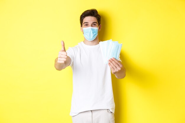 Concetto di covid-19, quarantena e misure preventive. Uomo soddisfatto che mostra pollice in su e dà maschere mediche, in piedi su sfondo giallo.