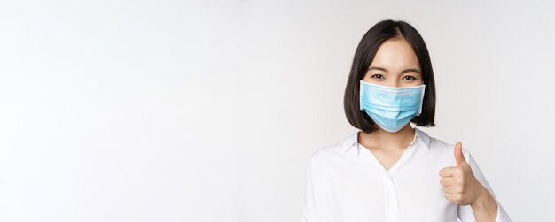 Concetto di Covid e assistenza sanitaria Ritratto di donna asiatica che indossa una maschera medica e mostra i pollici in piedi su sfondo bianco
