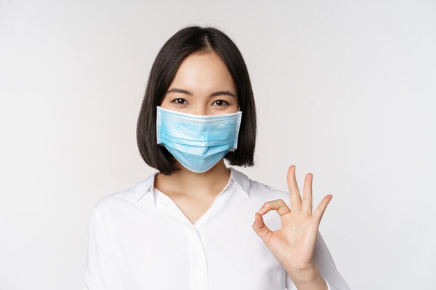 Concetto di Covid e salute Ritratto di donna asiatica che indossa una maschera medica e mostra un segno di ok in piedi su sfondo bianco