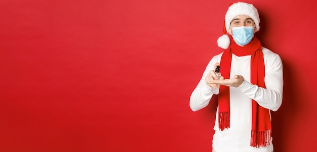 Concetto di covid19 natale e vacanze durante la pandemia Felice giovane uomo con cappello da Babbo Natale e maschera medica igienizza le mani con antisettico e sorridente in piedi su sfondo rosso