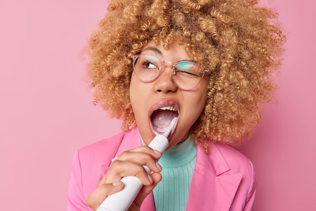 Concetto di cura dei denti e routine quotidiana La donna dai capelli ricci si lava i denti con lo spazzolino elettrico usa la pasta di menta si prende cura dell'alito fresco e dell'igiene vestita formalmente isolata su un muro rosa