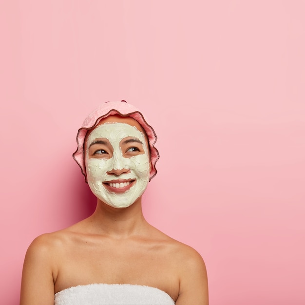 Concetto di cura della pelle professionale. La donna etnica felice applica la maschera facciale per pulire e idratare la pelle, guarda con un'espressione felice sognante, sorriso a trentadue denti, avvolto in un asciugamano morbido, pose al coperto