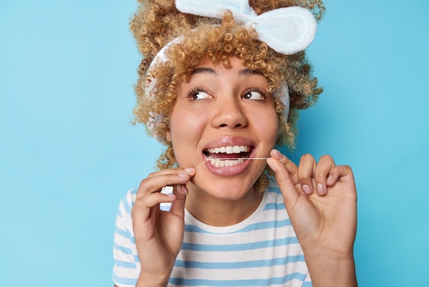 Concetto di cura dentale e carie dentaria La giovane donna riccia usa il filo interdentale indossa la maglietta a righe casual con fascia per capelli ha un sorriso perfetto distoglie lo sguardo isolato su sfondo blu studio