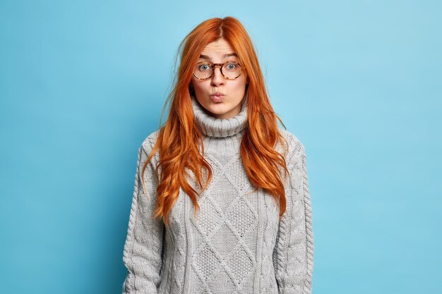 Concetto di espressioni facciali. La bella donna europea dai capelli rossi tiene le labbra piegate e sembra sorprendentemente che indossa un maglione lavorato a maglia.
