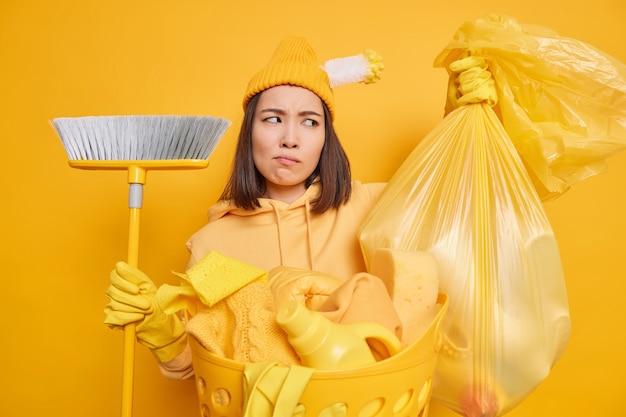 Concetto di faccende domestiche. La casalinga insoddisfatta che va a portare fuori la spazzatura dalla casa spazza il pavimento con le pose di scopa vicino al cesto pieno di biancheria e detersivi isolati su sfondo giallo