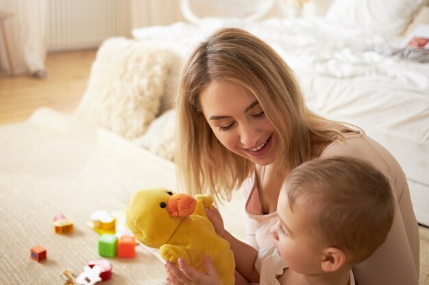 Concetto di famiglia, infanzia, maternità e prenting. Scena carina di mamma giovane bionda seduta sul pavimento in camera da letto con il suo adorabile figlio bambino circondato con giocattoli che giocano con anatra gialla farcita