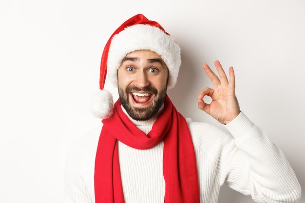 Concetto di festa di Capodanno e vacanze invernali. Primo piano dell'uomo attraente felice in cappello della santa che mostra il segno giusto, celebrante il natale, fondo bianco