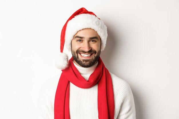 Concetto di festa di Capodanno e vacanze invernali. Primo piano dell'uomo caucasico allegro che celebra il Natale in cappello di Babbo Natale, sorridente felice, sfondo bianco.