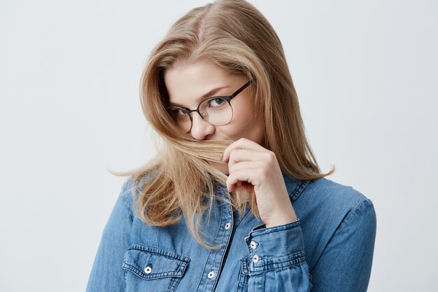 Concetto di gioventù e felicità. Bella ragazza adolescente caucasica in camicia di jeans, guardando la telecamera con appello, giocando con i capelli biondi. Giovane donna con una pelle sana perfetta in occhiali alla moda