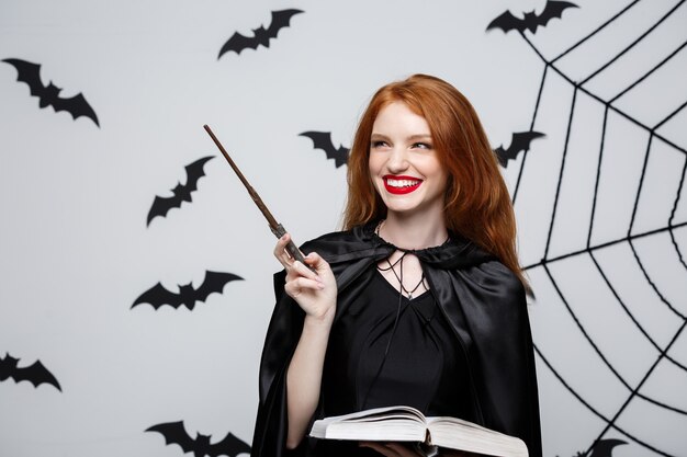 Concetto di Halloween - Bella strega che gioca con il bastone magico e il libro magico sul muro grigio.