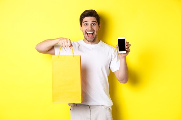 Concetto di mobile banking e cashback. Giovane ragazzo felice che tiene la borsa della spesa e che mostra lo schermo dello smartphone, sfondo giallo.