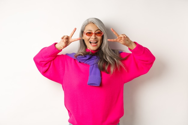 Concetto di moda e bellezza. Immagine di una donna anziana asiatica alla moda in occhiali da sole che sorride, mostra segni di pace e sembra felice, in piedi su sfondo bianco