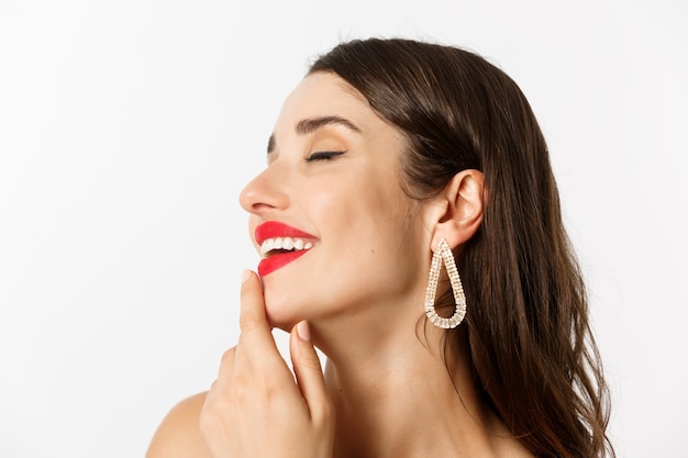 Concetto di moda e bellezza. Primo piano del volto di sensuale donna bruna con orecchini e rossetto rosso, sorridendo tentato e toccando il labbro, in piedi su sfondo bianco.