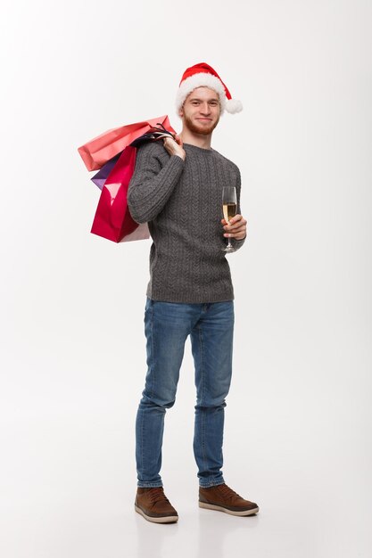 Concetto di Natale Giovane uomo con la barba bella che tiene un bicchiere di champagne e borse della spesa con felice espressione facciale