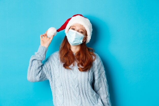 Concetto di Natale, quarantena e covid-19. Allegra ragazza dai capelli rossi con cappello da Babbo Natale e maschera facciale, fissando la telecamera compiaciuta, in piedi fiduciosa su sfondo blu.