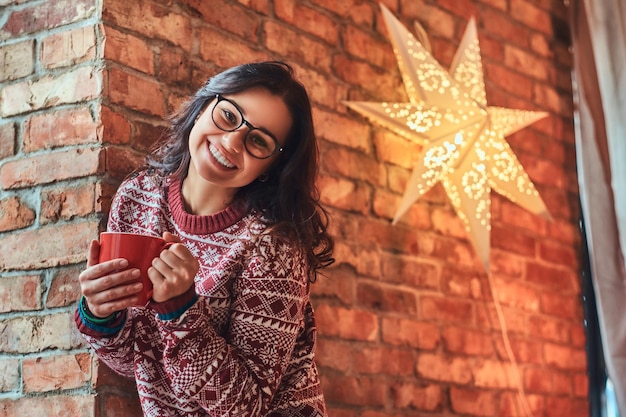 Concetto di Natale. Ritratto di una ragazza bruna allegra che indossa occhiali da vista e maglione caldo che tiene una tazza di caffè mentre si appoggia su un muro di mattoni.