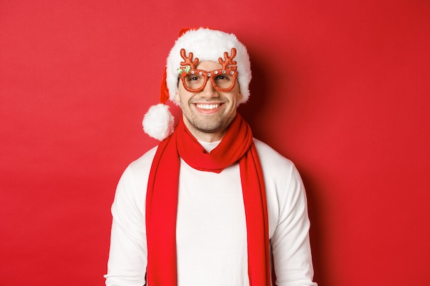 Concetto di natale, vacanze invernali e celebrazione. Bel ragazzo allegro con gli occhiali da festa di capodanno e sorridente, in piedi su sfondo rosso