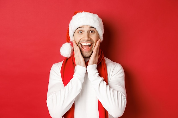 Concetto di natale, vacanze invernali e celebrazione. Primo piano dell'uomo sorpreso e felice in cappello e sciarpa della santa, guardando qualcosa di incredibile, in piedi su sfondo rosso.