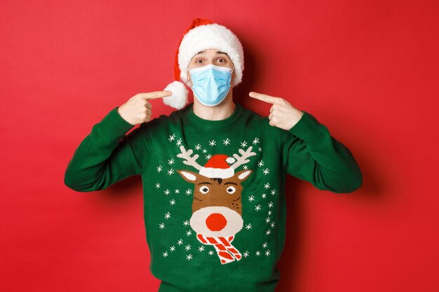 Concetto di nuovo anno, coronavirus e distanziamento sociale. Uomo felice con cappello da Babbo Natale e maglione natalizio, che consiglia di indossare una maschera medica alla festa, in piedi su sfondo rosso