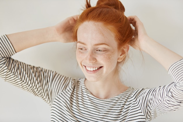 Concetto di persone e stile di vita. Attraente giovane donna con i capelli rossi e la pelle lentigginosa che indossa la parte superiore a strisce che sorride felicemente mentre regola la sua acconciatura prima di uscire a fare festa con i suoi amici