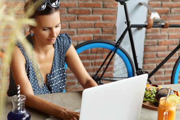 Concetto di persone e tecnologia moderna. Donna graziosa con le tonalità sulla sua testa che mangia pasto al caffè moderno con il muro di mattoni e la bicicletta rossi