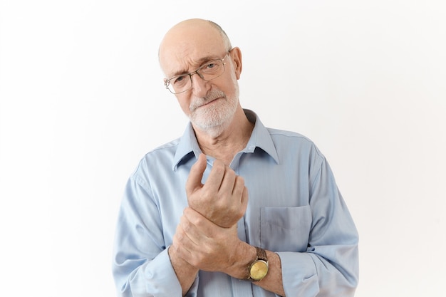 Concetto di persone, età, benessere, malattia e problemi di salute. Studio shot di frustrato sconvolto sessantenne uomo in occhiali con aspetto doloroso, sfregamento del polso, che soffrono di dolori alle articolazioni