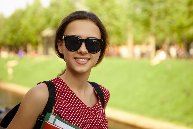 Concetto di persone, gioventù, scuola e istruzione. Ragazza del college positiva felice alla moda che indossa sfumature nere e zaino per il trasporto godendo del bel tempo estivo, tornando a casa dalle lezioni,