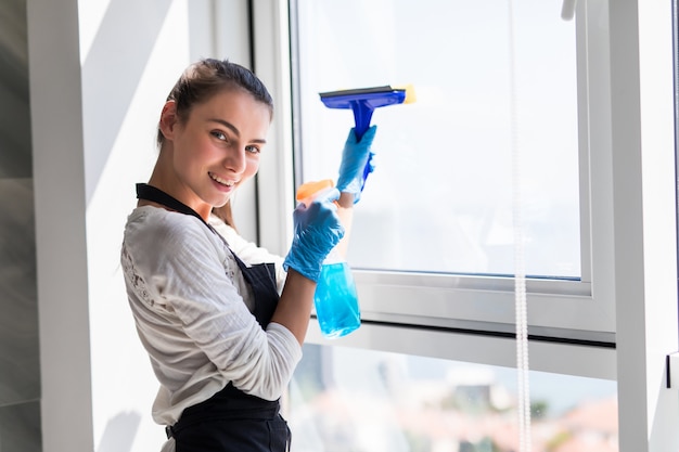 Concetto di persone, lavori domestici e pulizie. La donna felice in guanti che puliscono la finestra con lo straccio e la pulitrice spruzzano a casa