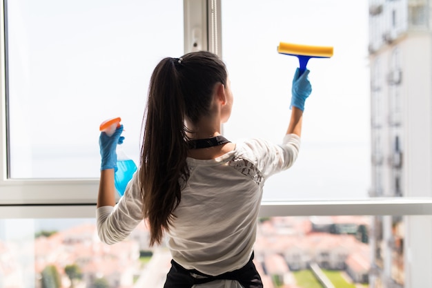 Concetto di persone, lavori domestici e pulizie. La donna felice in guanti che puliscono la finestra con lo straccio e la pulitrice spruzzano a casa