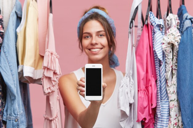 Concetto di persone, stile, moda, shopping e tecnologie moderne. Ritratto della donna allegra attraente nello spogliatoio o nel guardaroba che mostra telefono cellulare