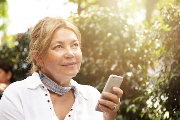 Concetto di persone, tecnologia e comunicazione. Donna senior affascinante con capelli biondi che per mezzo dello Smart Phone generico