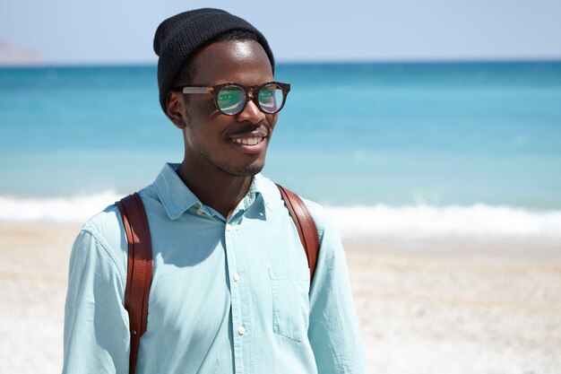 Concetto di persone, viaggi, vacanze e turismo. Giovane viaggiatore con zaino e sacco a pelo maschio dalla pelle scura allegro che gode della vista pittoresca e della vista sul mare durante le vacanze estive in stazione turistica, camminante sulla spiaggia da solo