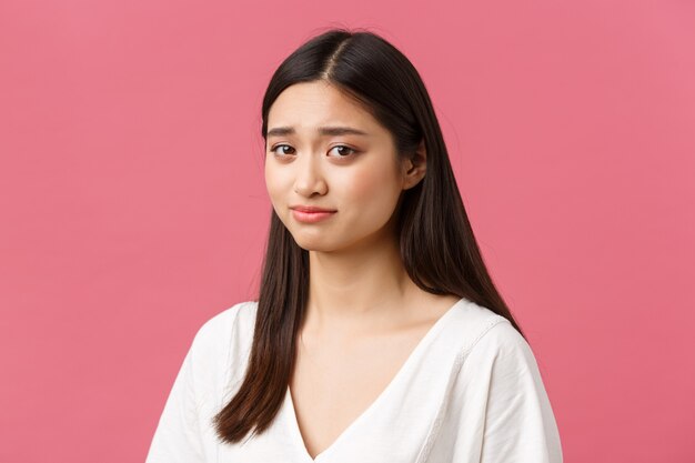 Concetto di pubblicità di prodotti per il salone di bellezza, la cura dei capelli e la cura della pelle. Primo piano di una ragazza asiatica carina scettica e insicura, sorrisetto imbarazzante, sensazione di disagio, sfondo rosa in piedi