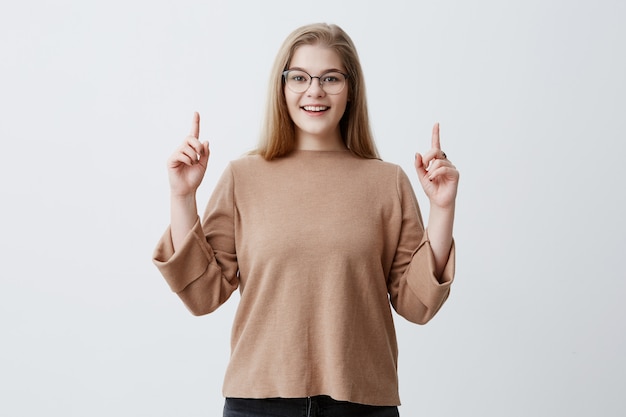 Concetto di pubblicità. La donna felice in maglione, indica con le dita anteriori come mostra qualcosa, ha sorpreso l'espressione compiaciuta. Femmina bionda che indossa i gesti alla moda degli occhiali contro la parete grigia