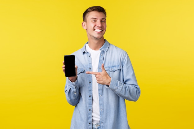 Concetto di pubblicità per il tempo libero, la tecnologia e l'applicazione. Ragazzo biondo sorridente felice e fiero che consiglia l'app per smartphone, puntando il dito contro il cellulare per vantarsi con le sue foto, sfondo giallo.