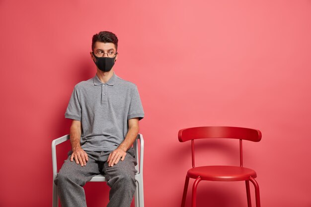 Concetto di quarantena automatica. Triste uomo solo indossa maschera protettiva rimane a casa durante l'isolamento essendo depresso a causa della situazione di focolaio si siede vicino a una sedia vuota