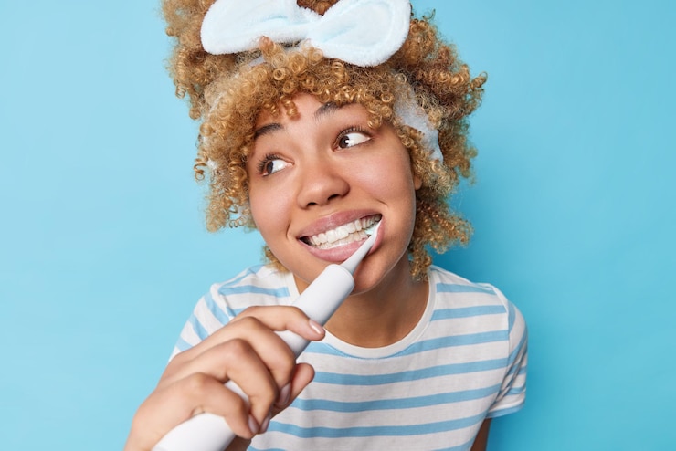Foto gratuito concetto di routine di igiene orale bella donna con capelli ricci pettinati distoglie lo sguardo spazzola i denti con uno spazzolino elettrico fa una cura dentale quotidiana vestita con una maglietta a righe isolata su sfondo blu