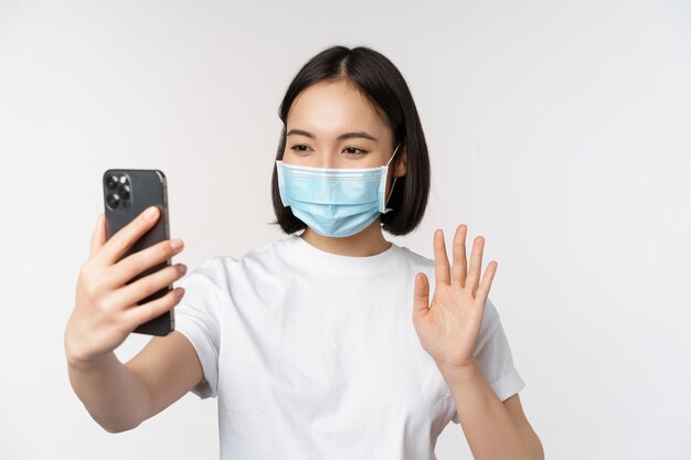 Concetto di salute e covid19 Studentessa asiatica moderna in video chat con maschera medica con telefono cellulare che agita la mano all'app per smartphone in piedi su sfondo bianco