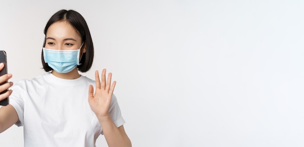 Concetto di salute e covid19 Studentessa asiatica moderna in video chat con maschera medica con telefono cellulare che agita la mano all'app per smartphone in piedi su sfondo bianco