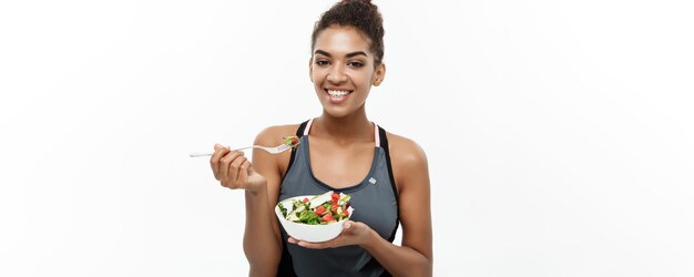 Concetto di salute e fitness Bella signora africana americana in abiti fitness a dieta che mangia insalata fresca isolata su sfondo bianco